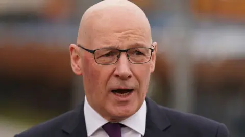 John Swinney, mid speech, wearing black spectacles, a black blazer, a white shirt and a purple tie. The shot captures him from the shoulders up.