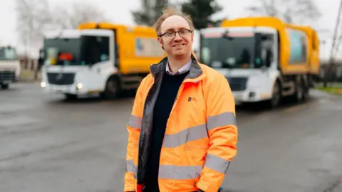 Hull City Council A man with short ginger hair and rectangular glasses is smiling into the camera. He is wearing a orange high-vis coat, a black jumper and shirt. Behind him are two recycling vehicles which are blurred.