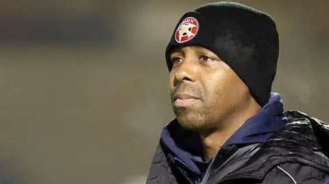 Darren Byfield stands on the touchline in his black Walsall beanie and club coat