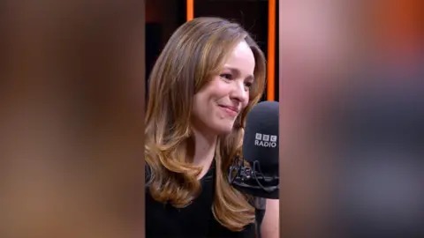 Rachel McAdams sits behind a BBC microphone, she has shoulder-length dark blonde hair and wears a black T-shirt