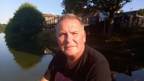 A man with short brown hair and a dark top sitting beside a pool of water with buildings and trees on the far bank.