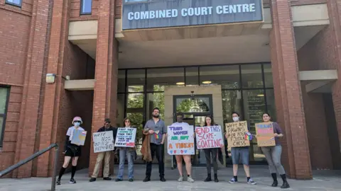 Submitted A group of people holding colourful signs which read messages such as 'trans women are not a threat', 'end this scape-goating' and 'we're not pawns for your political chess'. They are standing at the top of stairs outside a court which says 'Combined Court Centre' at the top.