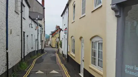 Silver Street in Honiton, Devon. It is a narrow single lane one-way road with buildings either side of the road. Double yellow lines are running alongside both sides of the road.