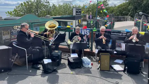 BBC / Dave Edwards Five people sitting in chairs outside playing musical instruments. They are looking at sheets of music on stands with speakers in front of them. In the background there are railings which are decorated with colourful bunting.