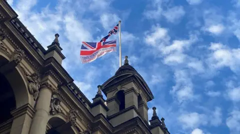 BBC Two flags coloured red, white and blue attached top one white flag pole on top of a stone tower with blue sky in the background