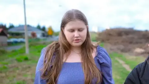 BBC Julia and Antillet are walking across a field with her eyes down. She wears a purple dress and has long -brown mouse hair.