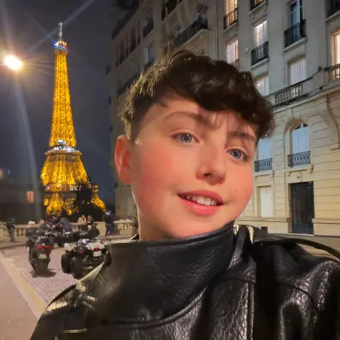 Harry Sawtell A teenage boy with brown hair takes a selfie in front of the Eiffel Tower at night