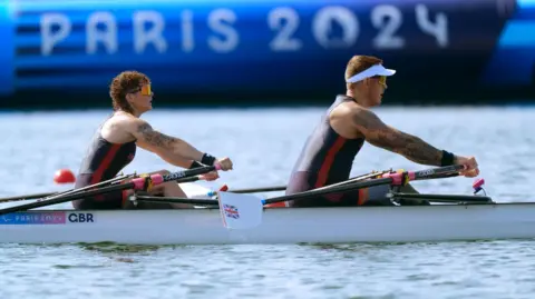 PA Media A man and a woman rowing a white boat. The woman sits at the back and has curly brown hair. The man sits at the front and has short blonde hair. They are both wearing sunglasses, and black and red wetsuits.