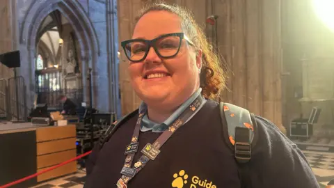 A woman wearing thick framed glasses and smiling. Her curly hair is in a ponytail. She is wearing a blue jumper that says "guide dogs" in yellow print. She also has a lanyard with different badges pinned on.