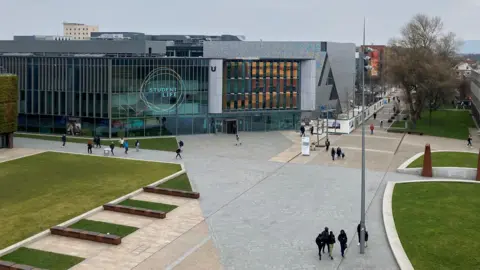 Teesside University, viewed from a height, with glass fronted buildings surrounded by concrete pathways interspersed with some curved lawns and a section of rectangular lawn. Students - looking small because they're down below - are walking about. One of the glass frontages - which look dark grey from outside - has a sign saying Student Life on it in white lettering.