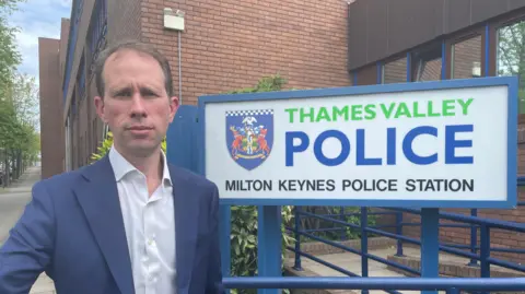 Matthew Barber has short brown hair and is wearing a white shirt and blue blazer. He is stood in front of Thames Valley Police's Milton Keynes station.