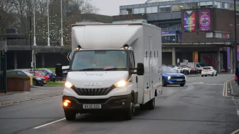 PA Media A white prison van travelling down a road in Swindon with its headlights on.