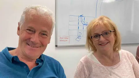 A man in a blue shirt sits next to a woman in a pink top with glasses and both are smiling and looking happy.