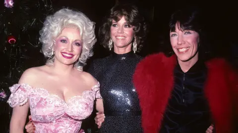 Tom Wargacki/WireImage Dolly Parton, wearing a bright pink off-the-shoulder dress, Jane Fonda in a black sequinned high neck dress, and Lily Tomlin in a black dress and bright red fur wrap.