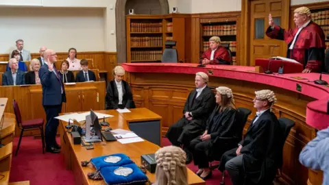 PA Media John Swinney stands with his right hand raised in a court room, facing the lord president, who is wearing a red robe and wig. The lord president is standing, also with his right hand raised, with court officials looking on 