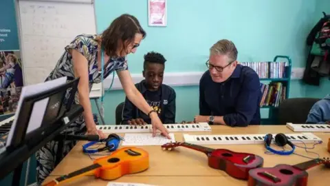Reuters The prime minister sits at a table with guitars and headphones on it. A young boy sits next to him and a woman stands on the boy's other side, pointing at a sheet of music.