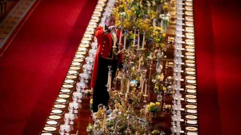 PA Media A member of Royal Household staff lights a candle during table preparations in St George's Hall, they wear a red jacket and black trousers. the long table is decorated in lavish floral arrangements and candle sticks. A row of plates and cuttlery line the edges of the table.