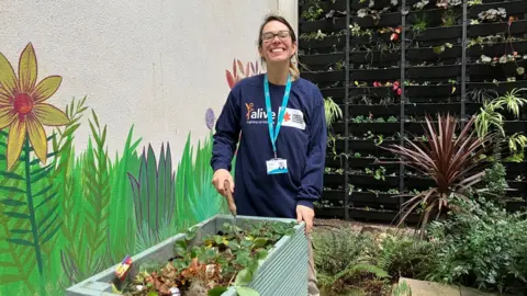 Abi Sweet from the charity Alive holds a gardening tool and stands at a tall light blue vegetable planter in front of a living wall that features a variety of small plants.