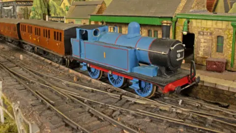 Talyllyn Railway collection A close up picture of part of a model railway layout with a bright blue steam engine on the track, pulling two orange-brown carriages. The locomotive has a number "1" on the side in the style of Thomas the Tank Engine, but does not have a face. A model station is in the background.