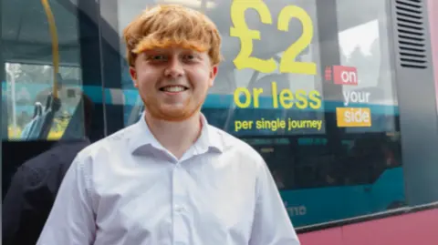 A man with ginger hair and a ginger beard stands next to a red bus while wearing a white shirt. He is smiling at the camera with his hands together.