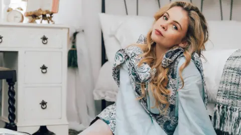 Bryony Coles Saoirse-Monica Jackson has long blonde curly hair. She is wearing a flowing light blue blouse.