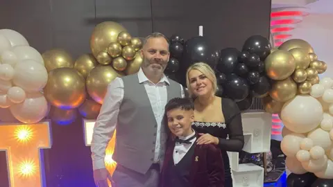 Children with Cancer UK A smartly dressed man, woman and boy are smiling together at the camera and standing in front of a cluster of white, gold and black balloons