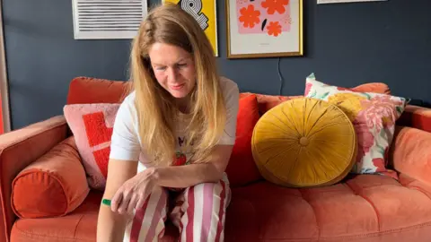 Rachel Verney A woman with dark ginger hair is wearing a white t-shirt and is sitting on a bright orange sofa.
