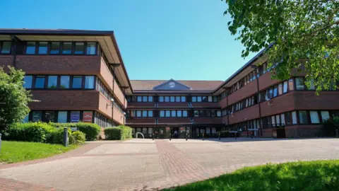 Gateshead Council A large brick building with three floors and multiple windows. There is a clock above the entrance. There is a block paved driveway and grass and bushes n bedding areas.