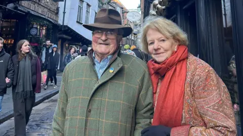 Joanna Spicer Joanna and Christopher standing in Abbeygate Street in Bury St Edmunds. Christopher wears a green tweed jacket and brown trilby and glasses. Joanna stands to his left, leaning towards him in her red patterned coat and scarf.