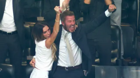 Victoria Beckham throws her arm up in a celebratory fist as she hugs her elated husband following England's goal against Argentina on Thursday night