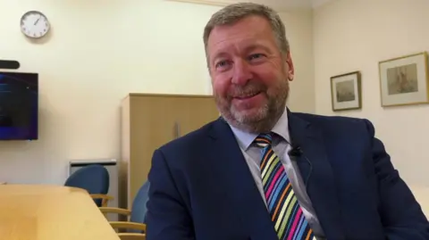 Sean Matthews wears a dark blue suit with a bright stripy tie and sits at a desk in a meeting room.