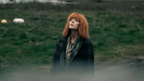 Kelly Reilly standing in a field, looking to the sky with her eyes closed. She has copper shoulder length hair with a fringe. She wears a green shirt and a green tartan coat.
