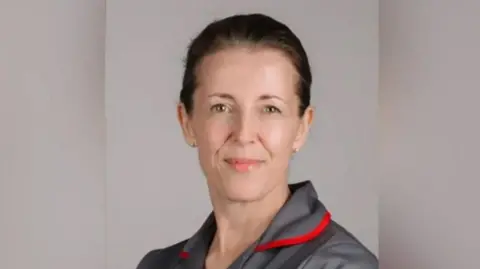 RJAH A woman in a grey nursing uniform smiling at the camera. She has dark brown hair pulled back and wears grey studs in her ears. there is red piping around the uniform's collar.