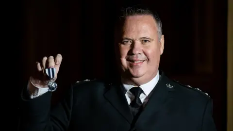 PA Media John Caldwell holds up his King's Police Medal while smiling at the camera.