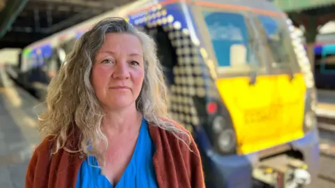 On the left, Rachel looks into the camera. She has grey wavy hair, and is wearing a mid-blue top underneath a copper-brown cardigan. In the background on her right there is a blue and yellow ScotRail train.