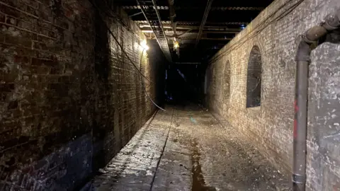 A dark tunnel with cobbled ground. On the right is the original station wall while on the left is the newer external wall built when the station was expanded.