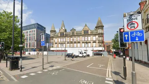 Google A large paved area with trees on one side and a boarded up yellow brick building which has a sign in red lettering which reads TJ Hughes. A white road sign with black writing which reads pedestrian zone and a blue road sign showing a cycle lane and an image of a bicycle and arrows are next to a set of traffic lights.