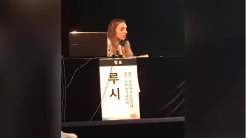 Lucy Robinson Lucy Robinson speaking at an event. Her name and other text in Korean are displayed in front of her.