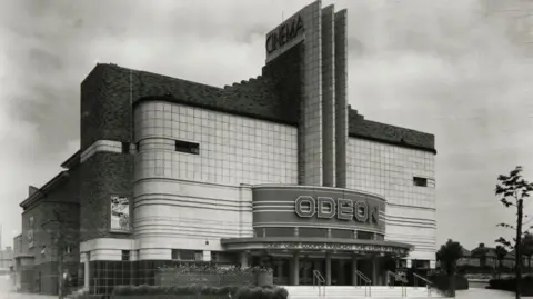 Historic England Archive An Odeon building which has an Art Deco structure with rounded corners and horizontal lines. There is a central tower rising above the entrance with horizontol straight lines. At the bottom “ODEON” is displayed. There are steps leading up to the doors. 