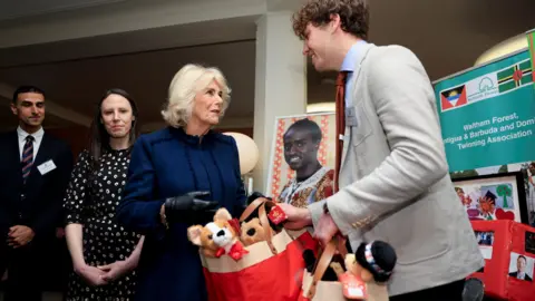 PA Media Queen Camilla, wearing a dark blue dress, hands over a Christmas-red bag containing toy stuffed animals to a representative of Citizens UK, as two people look on.  