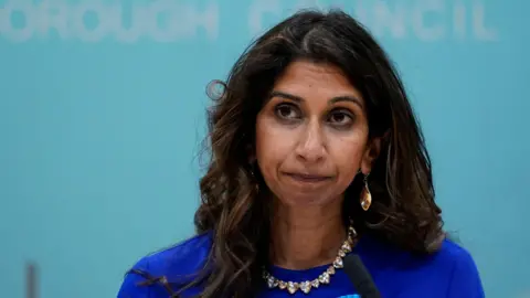 PA Media Suella Braverman MP is pictured with a serious expression in a council room. She has wavy, mid-length, dark hair and wears a blue top, jewelled necklace and earrings.