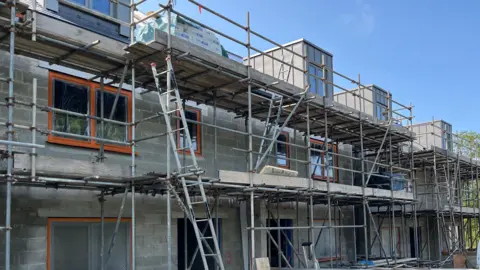 Scaffolding is in front of a row of new homes. The shell of the houses has been been with grey walls and brown edged window frames.