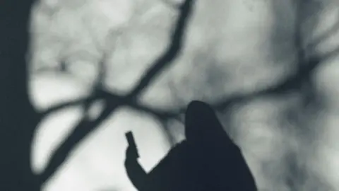 Milamai/Getty A greyed out silhouette of a person holding a phone against a blurred grey background, seemingly featuring skeletal trees, again in silhouette