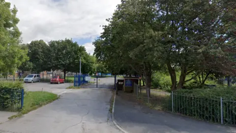 Google An exterior view of the entrance to Mill Rythe Infant School, there are primary blue coloured gates on a road, with a car park to the left which has a red fiat parked and a silver people carrier, there are trees lining either side of the road