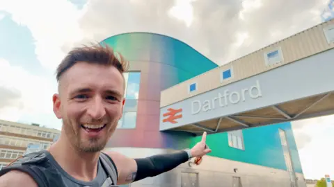 Matt Nelson A man, Matt Nelson, wearing running clothing and pointing at a sign behind him on a rail station tunnel. The sign says "Dartford" and has a railway symbol next to it.