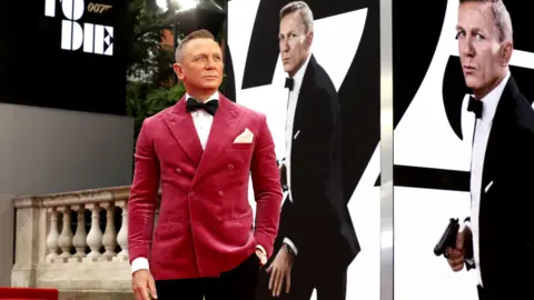 Daniel Craig appears at the premiere of No Time to Die (2021) wearing a striking berry suit jacket and posing in front of pictures of himself playing James Bond