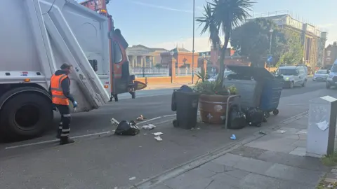 Phil Harrison/BBC A binman is collecting waste on a residential street. The bins are standing near the kerb and there is a black bin bag of waste on the ground. There are buildings and cars in the distance.
