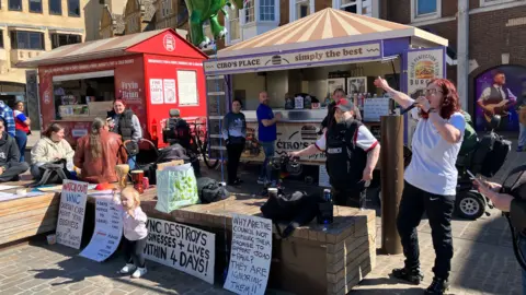 Sam Read/BBC Several people protest next to two food vans. There is a table with placards on it. The van is branded CIRO'S PLACE - SIMPLY THE BEST. It is a sunny day.