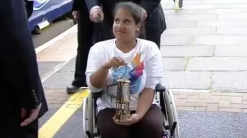 Doaa has her dark hair tied back. She is wearing a white shirt and red trousers. She is seated in a wheelchair and is carrying a lit lamp