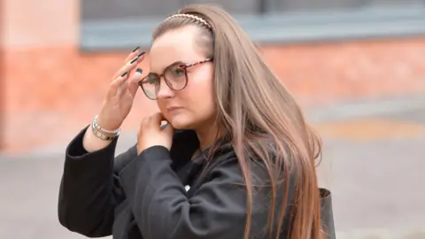 Spindrift A woman with long brown hair, a headband and glasses walking along a street. She is photographed from the chest up and is wearing a black coat and carrying a black bag. 